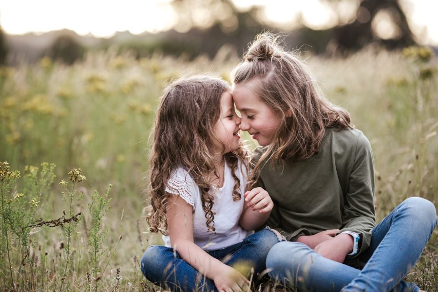 Two young girls are sitting cross legged in some long wild grass they have their faces and noses close together.