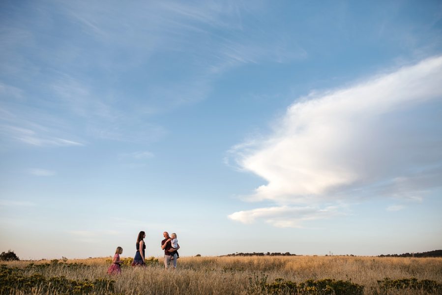 A family in the distance against an epic blue sky with interesting white clouds, they are slightly separated from eachother but the father is holding the youngest child.