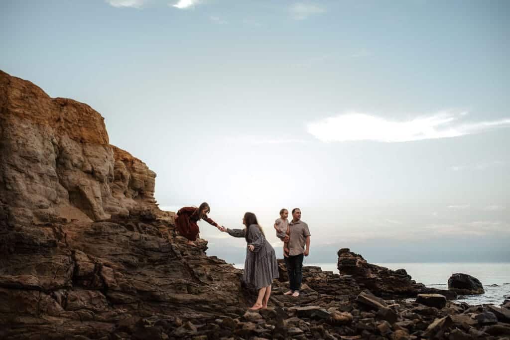 a girl reaches to her mother as her family stand on the beach rocks