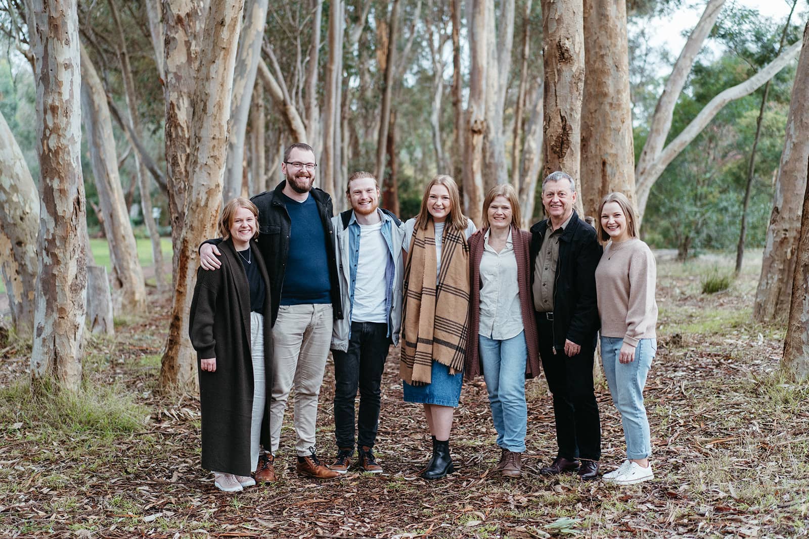 an extended family of parents and adult children look at the camera in a row they look comfortable and are in amongst striking gum trees
