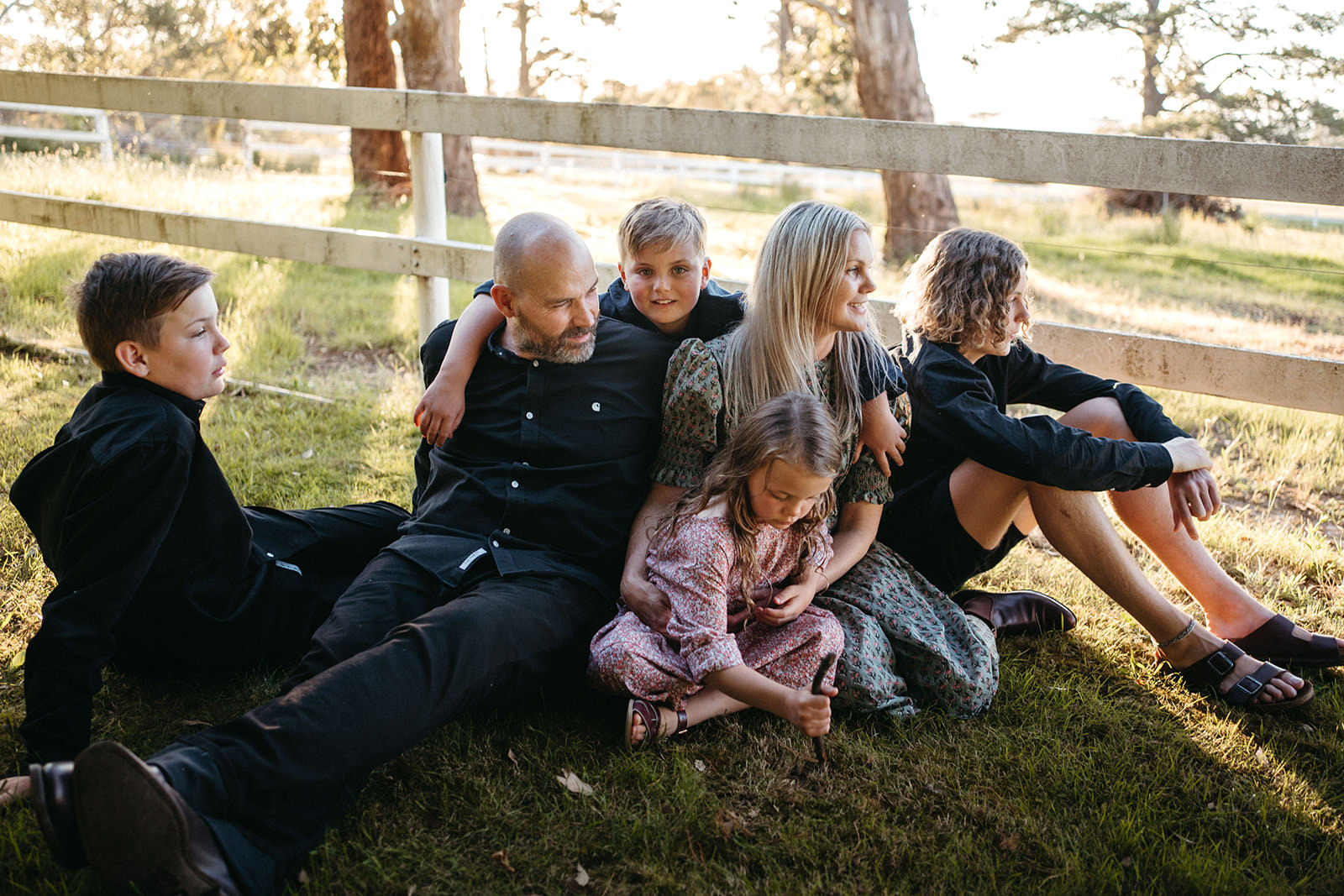a family of six sits on the grass in front of a fence outdoors, they are casual but connected and only one child is looking at the camera