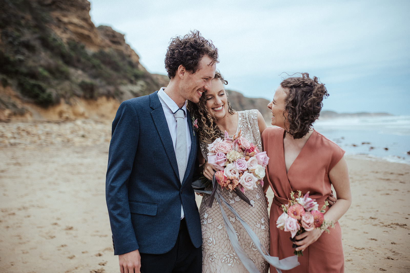 a bride hugs her brother and sister they are on the beach, both women are holding bouquets of pink and white flowers, the bride is leaning into her brother