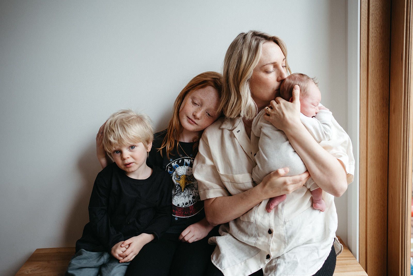 A mother sits with her three children she is kissing the youngest, they are sitting close and leaning together