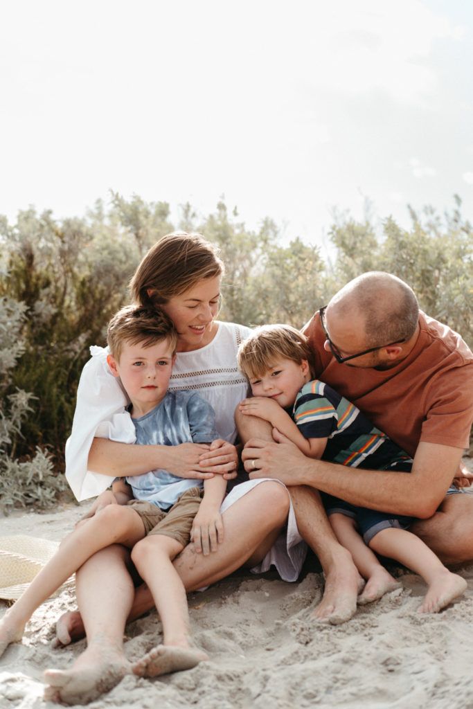 a family of mother father and two young boys cuddle together on the beach