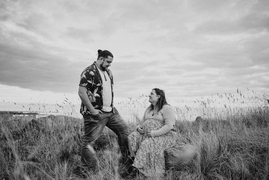 a pregnant woman looks up at her partner she is sitting on rocks in the grass at a Williamstown beach