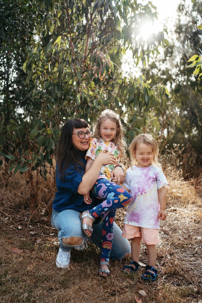 a mother squats on the ground with her two children (faces pixellated) they are wearing bright clothes.