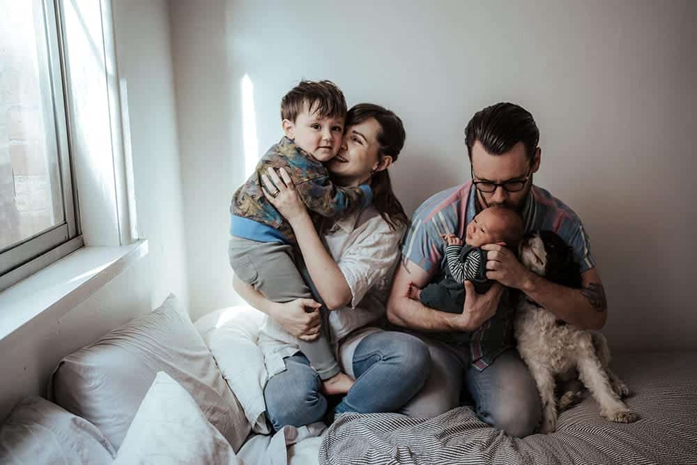 a family of four with a dog on a bed with a young toddler and baby they are hugging and dad is kissing newborn baby