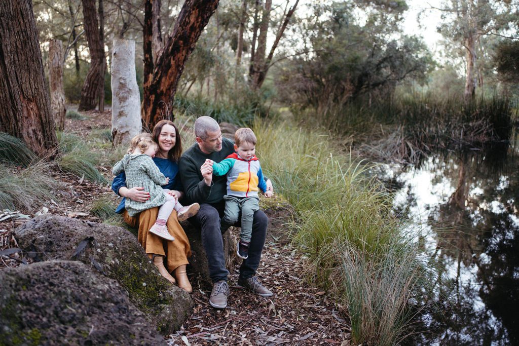 a family near a bushland pond sit together wearing bright primary colours