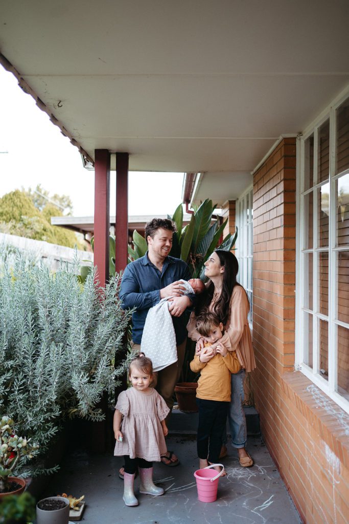 a family stand at the front of their house they are wearing plain colours but different colours with a pink bucket and chalk on the ground in front of them