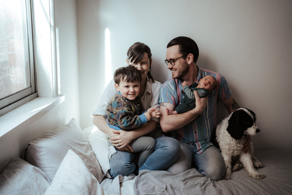 a family portrait of a mother and father and young boy and baby on a bed with a black and white dog. The mother is laughing