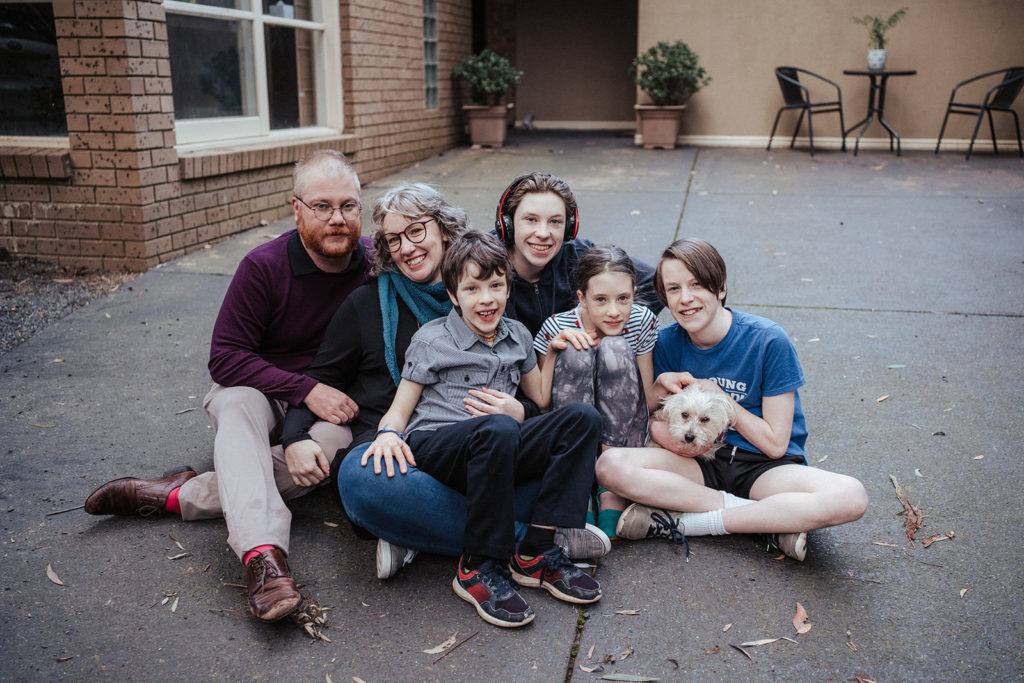a family portrait of six people sit on a driveway with a white dog, the children are older and one is neurodivergent and wearing headphones