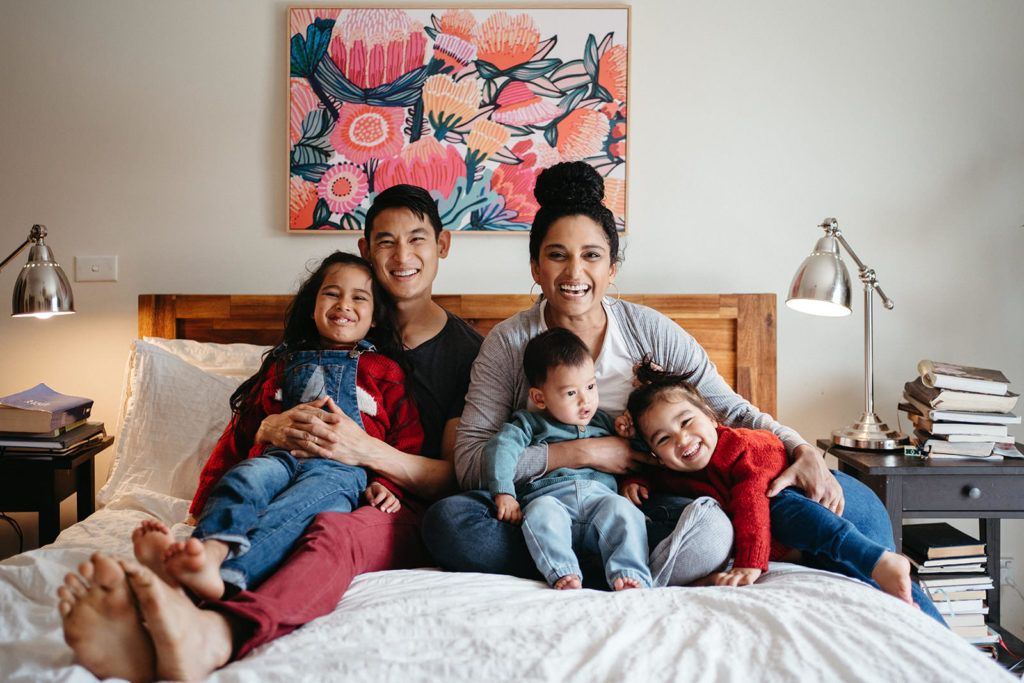 a family portrait of an Australian mixed race family with three children sitting on a bed with a bright pink floral artwork behind them, they are laughing and smiling at the camera