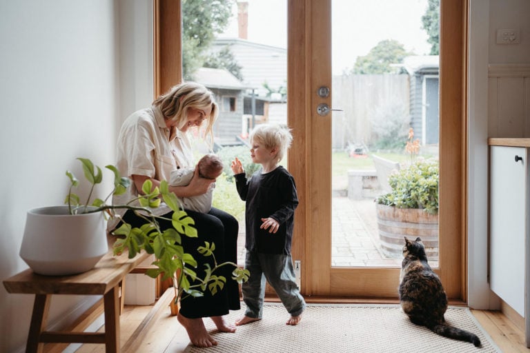 a mother sits holding her newborn son and her toddler reaches out a hand to touch his head a cat sits looking out the window