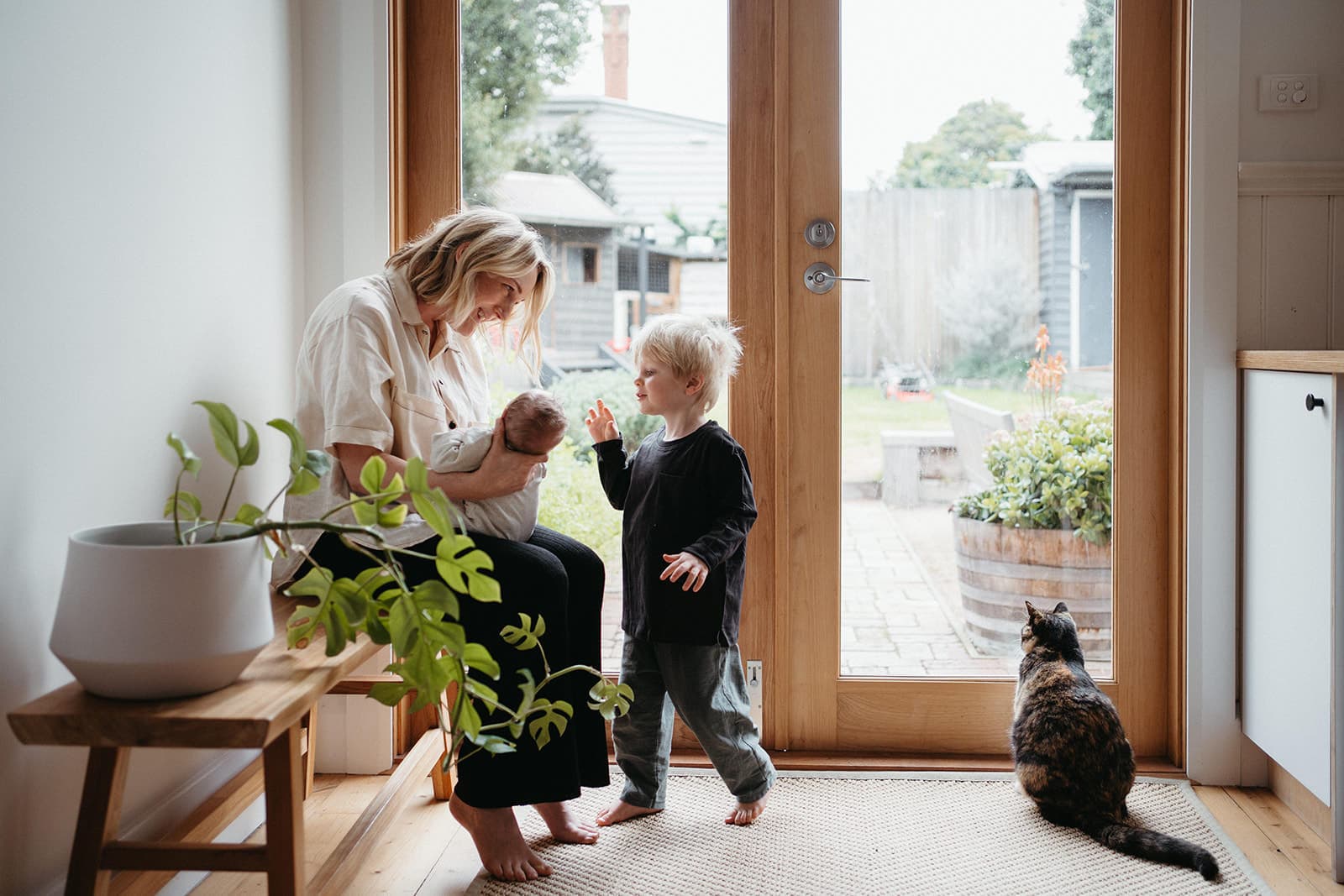 a mother sits holding her newborn son and her toddler reaches out a hand to touch his head a cat sits looking out the window