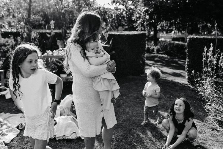a documentary image by a melbourne family photographer of a mother looking back at two children while one looks back at her, the mother is holding a baby