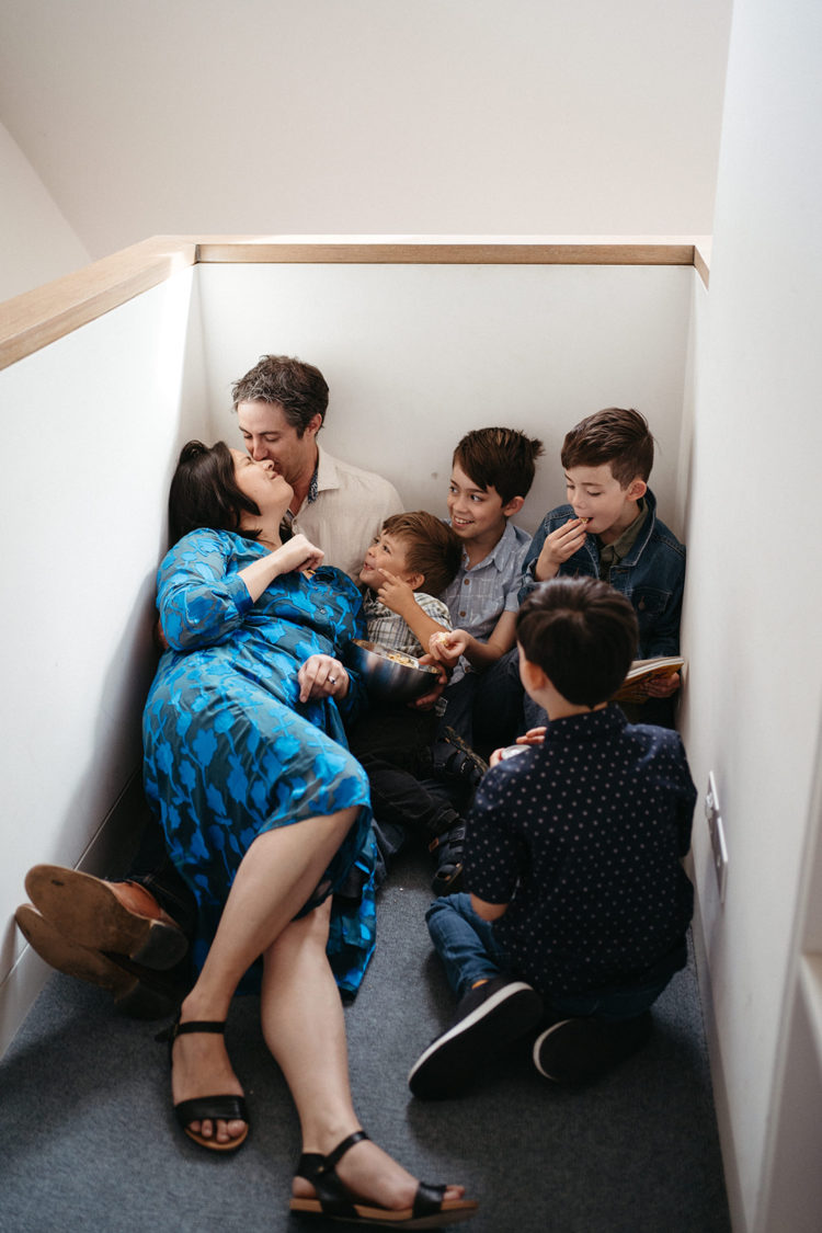 A family sits in a stairwell snuggled up the mum is wearing a blue dress and casually leaning back on the dad, four boys who are eating popcorn look on happily