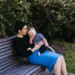 a woman in a black top and bright blue skirt sits on a park bench in Melbourne holding her young son