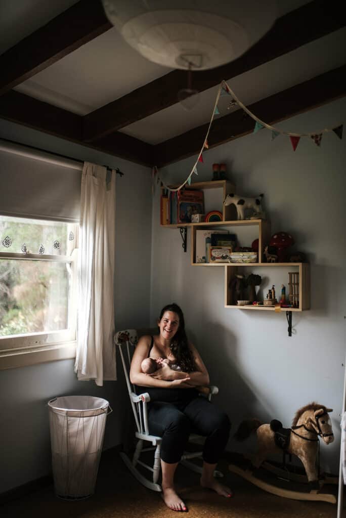 a woman sits in a chair breastfeeding her newborn baby she is looking at the camera and smiling