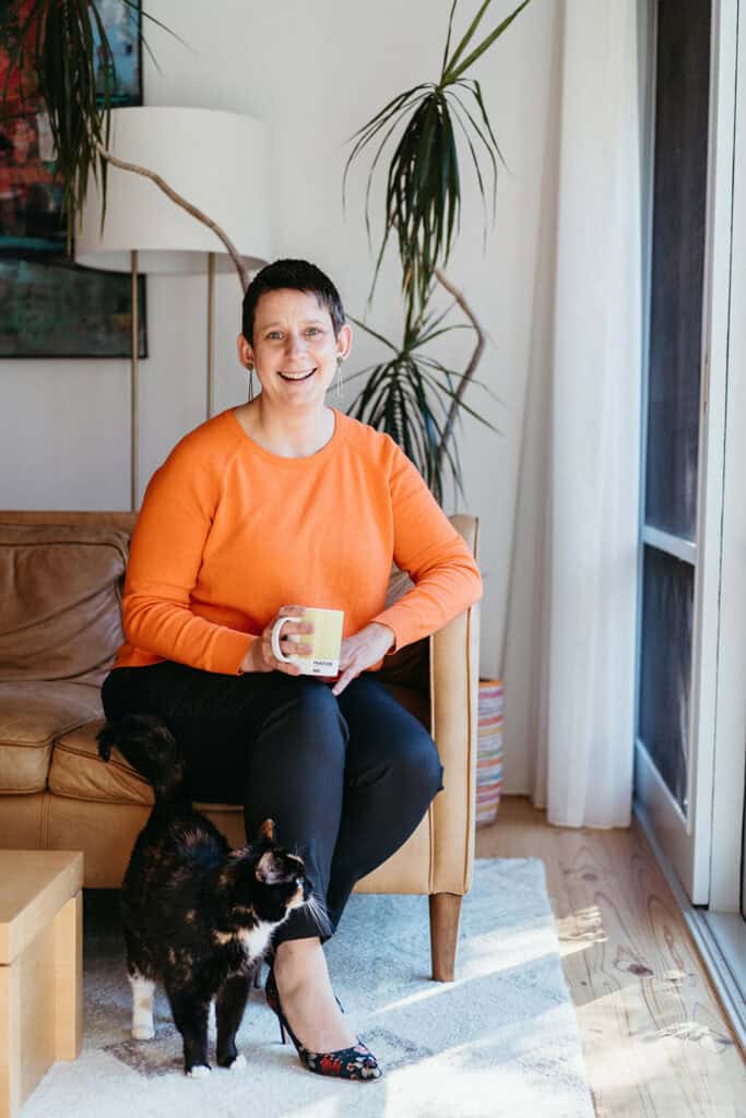 A woman in an orange top poses for an informal business portrait with a cat at her feet