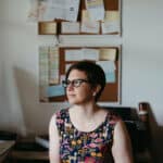 a portrait of a copywriter sitting at a desk with a wall of post it notes behind her, she is wearing glasses and a colourful dress
