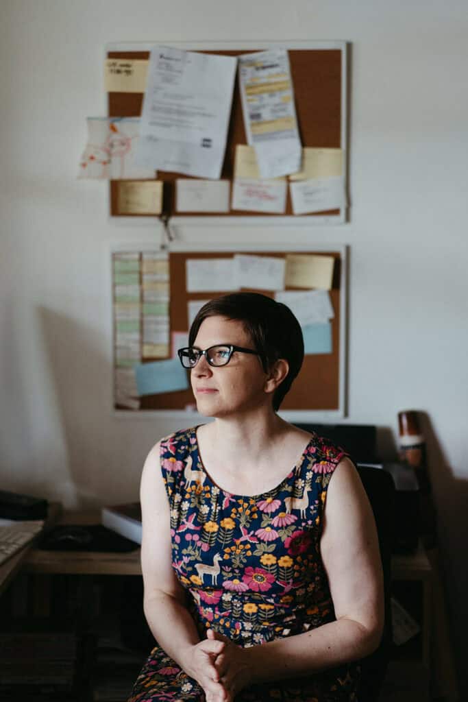 a portrait of a copywriter sitting at a desk with a wall of post it notes behind her, she is wearing glasses and a colourful dress