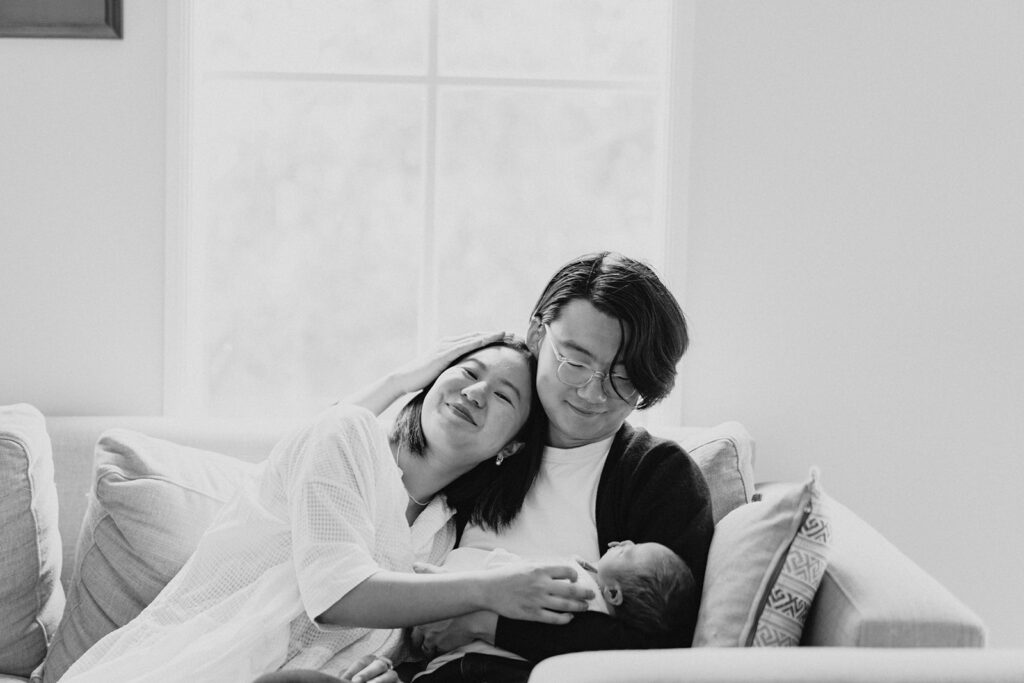a man holds his newborn baby and his wife leans in on him smiling he is reaching up to her and holding her closely