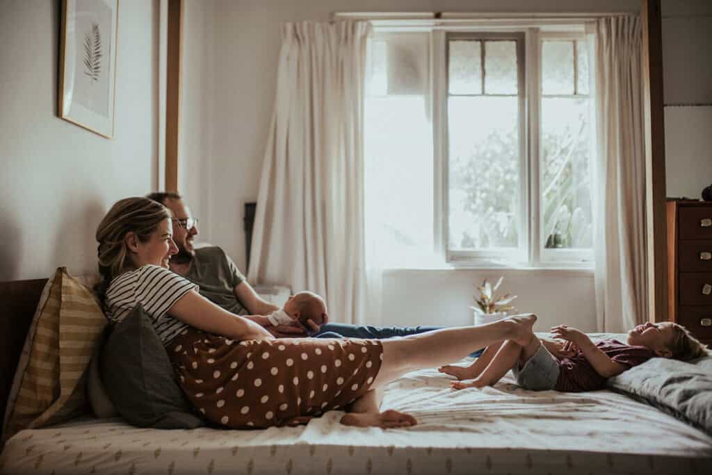 a mother reaches out with her foot to tickle her toddler who is giggling on a bed