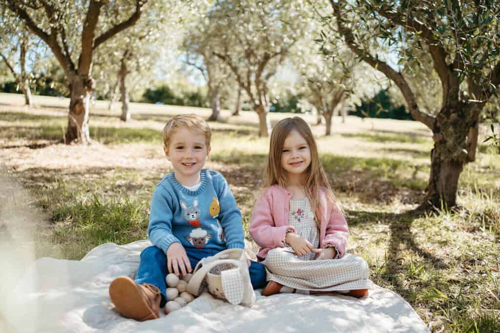 Two children sit on a rug in an Olive Grove wearing Purebaby Clothing