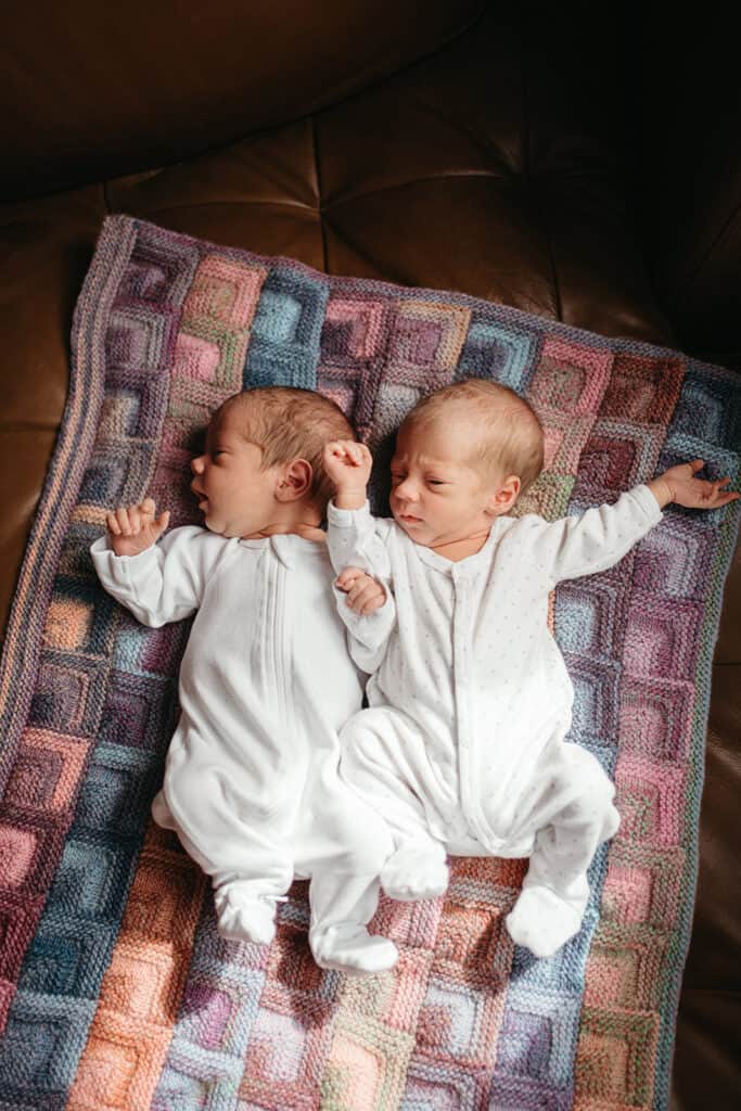 two baby twins in white onesies lie on a hand knitted blanket