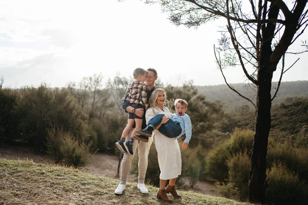 A family in Melbourne have their photo taken up a bushland hill the dad holds one boy and the mum holds another boy scooped up, they are smiling