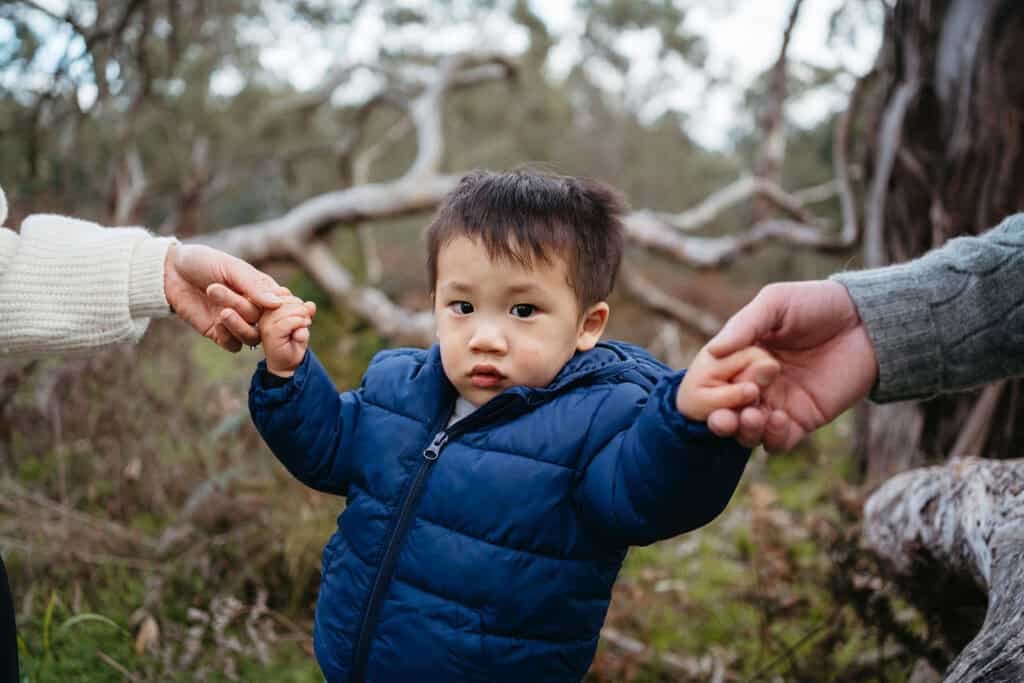 a young asian boy dressed in blue holds his parents hands during a photoshoot in the Australian bushland like in Bluey