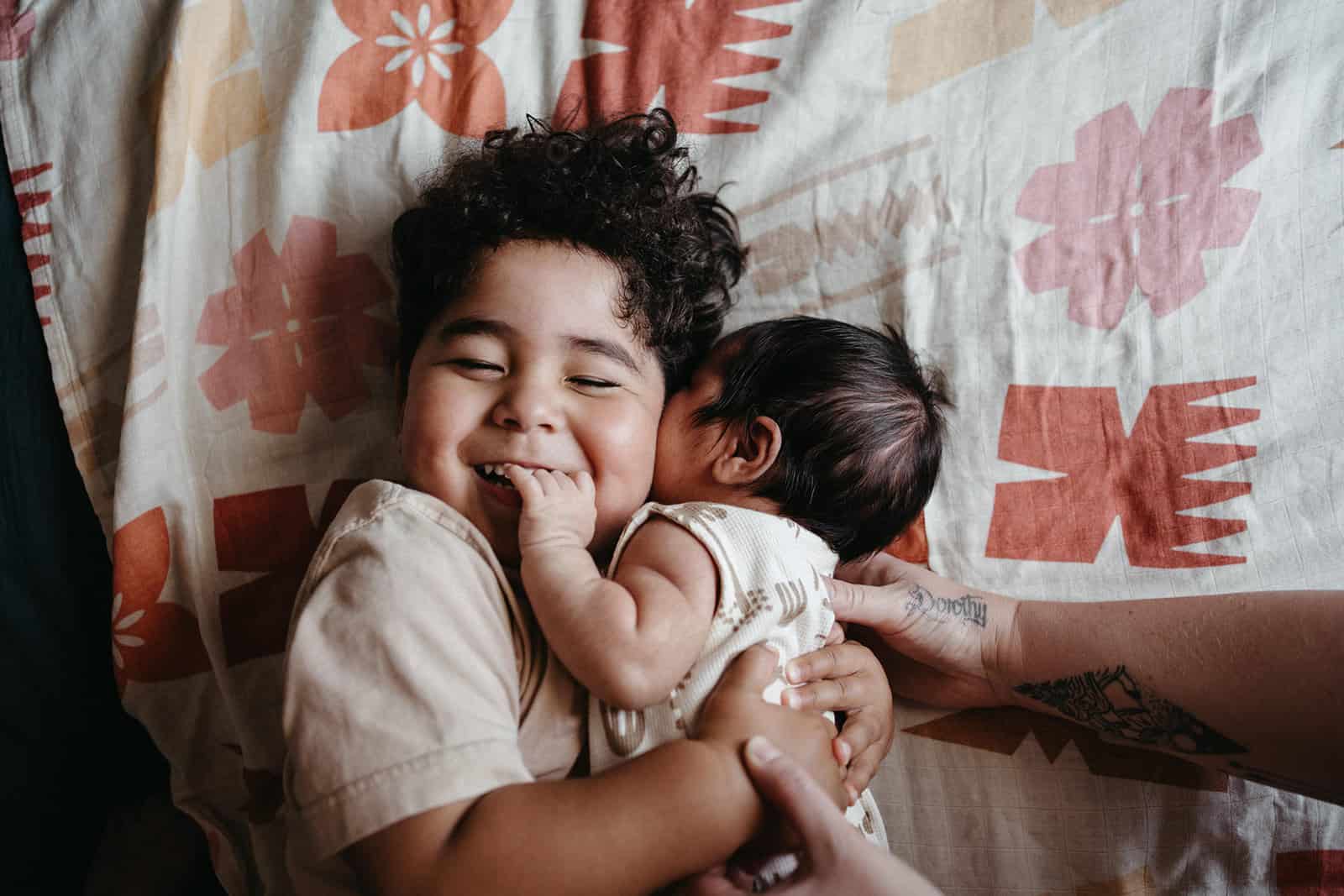 Toddler hugging baby brother during natural newborn photography session in Melbourne, lying on patterned island-style bedding