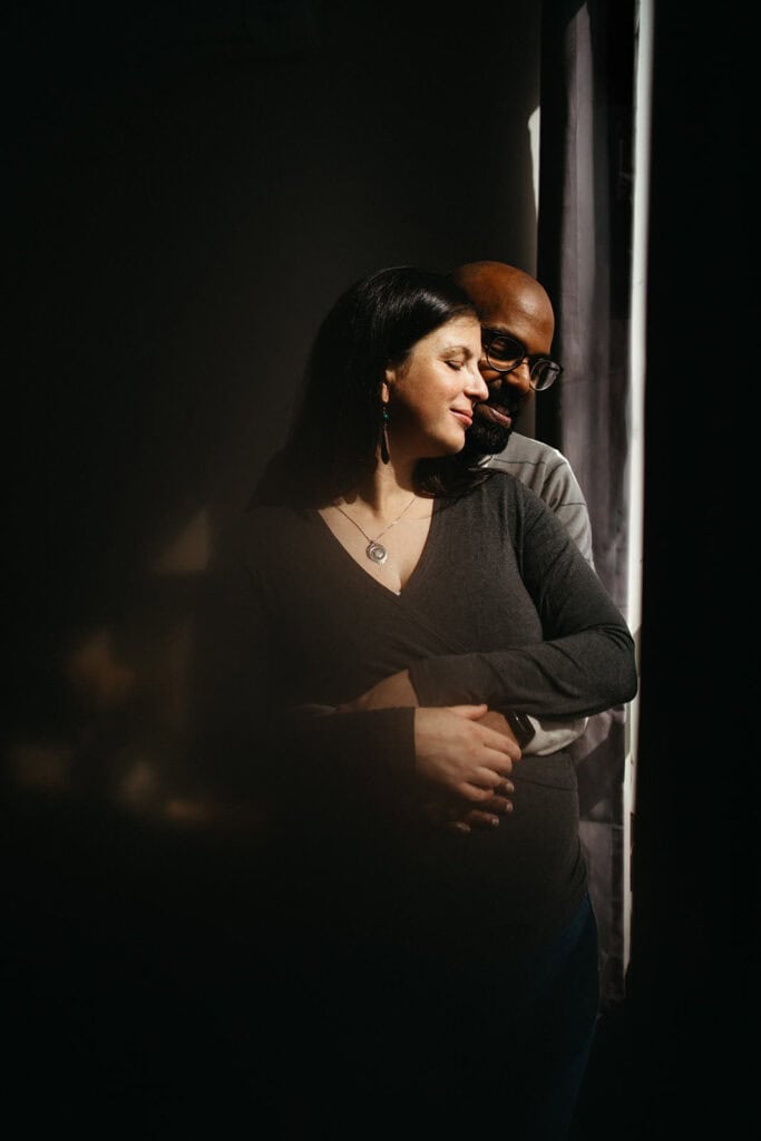 a pregnant couple stand near a window at a maternity session
