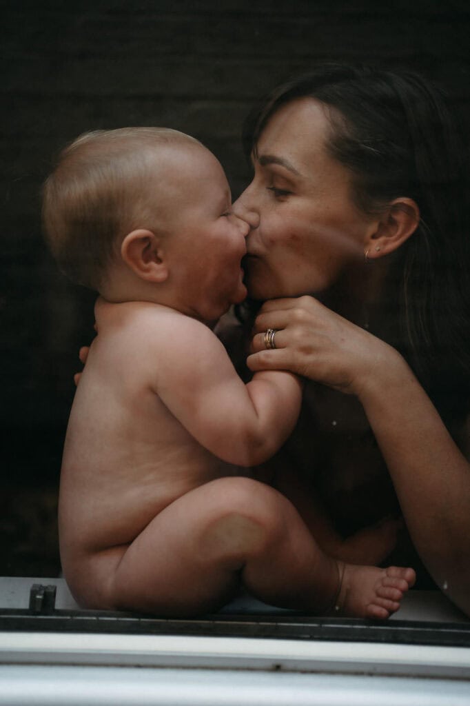 a mother kisses her chunky eight month old baby in a window image taken behind glass