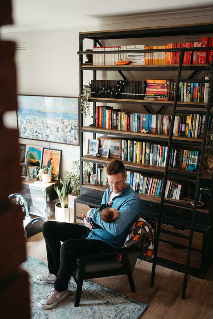 a man in a blue shirt holds his newborn baby infront of a colourful bookcase in a Melbourne home
