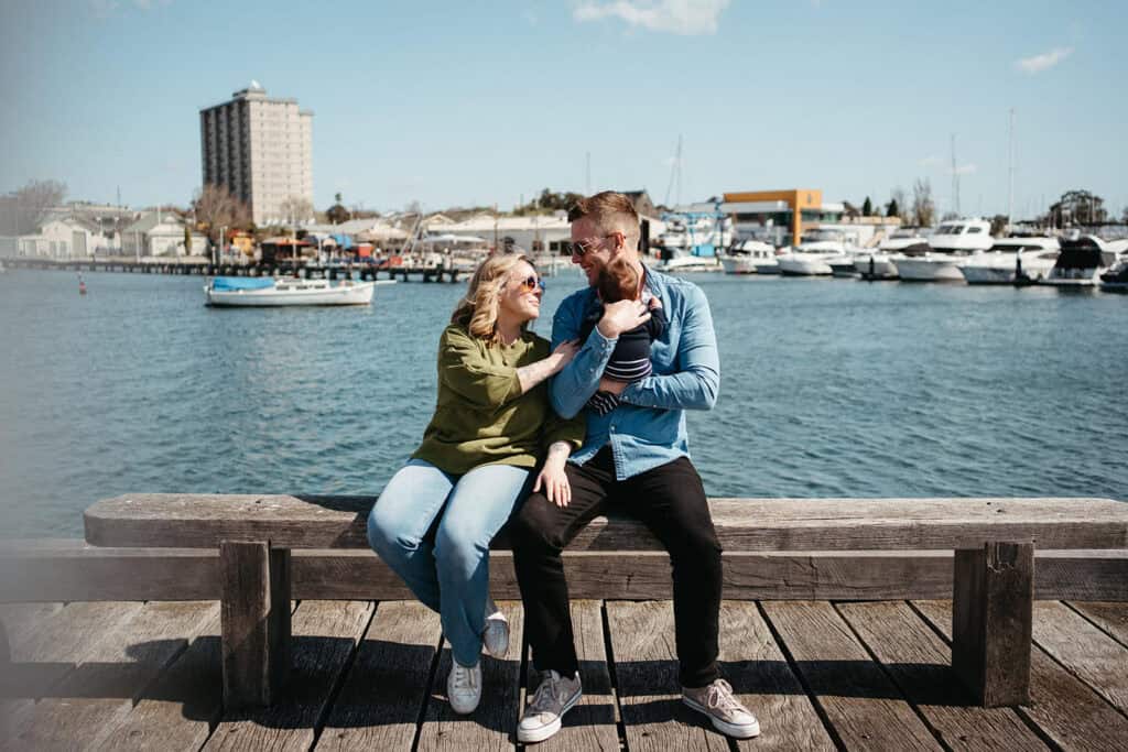 a mother and father wearing sunglasses sit on a bench in williamstown holding their new baby on a sunny day