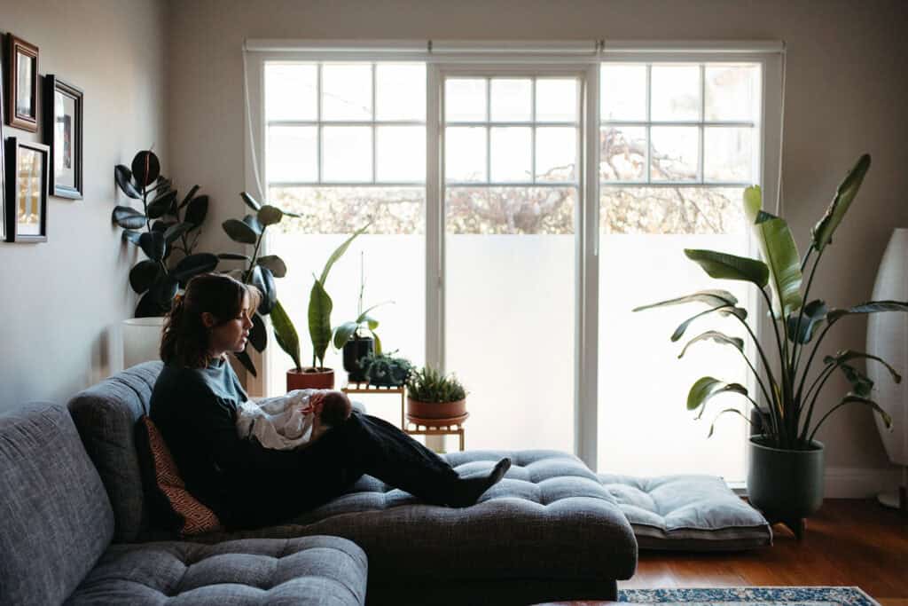 a mother sits at a window on a couch with her baby up on her knees