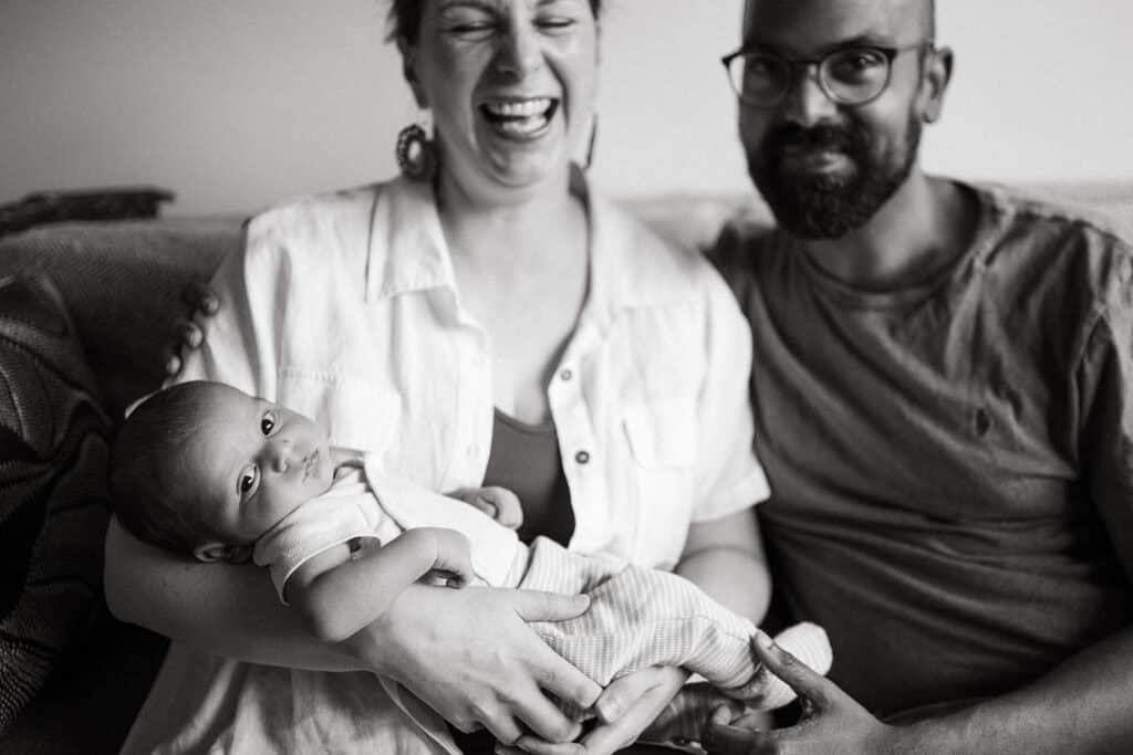 a black and white image of a mother laughing sitting next to her partner she is holding a baby who is looking directly at the camera