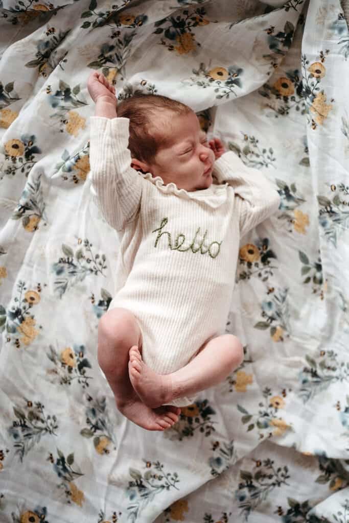 a newborn girl stretches she is wearing a onesie with an embroidered word hello on it she is on a floral muslin wrap