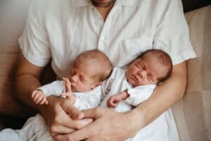 a father in a white shirt holds newborn twins in his arms they are at home