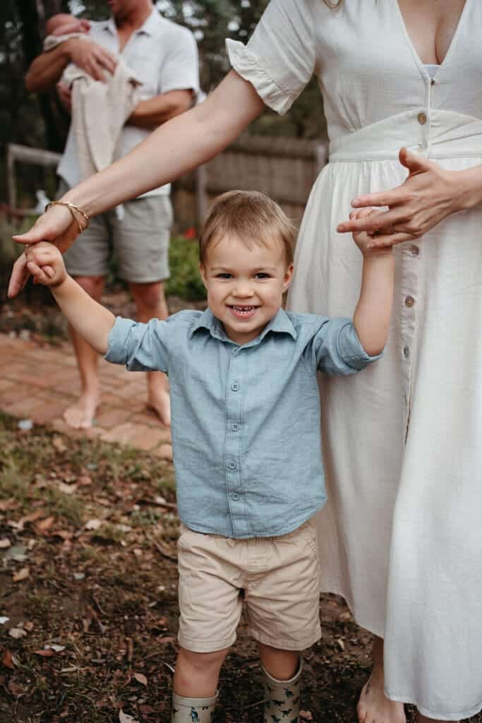 a toddler in the foreground of a newborn family photo session he is wearing a blue shirt and holding his mums hands she is in a neutral cream dress