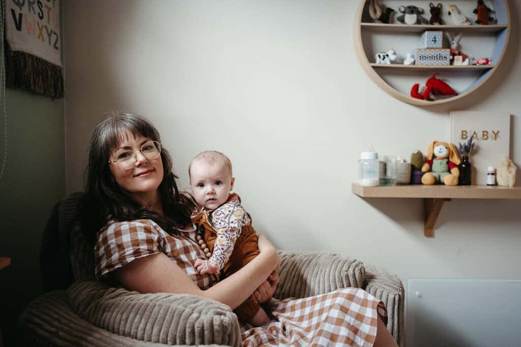 a mother in gingham is sitting on a nursery chair, she holds her three month old baby they are looking at the camera