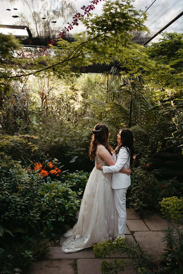a bride and bride stand in alowyn gardens greenhouse on their wedding day - they are looking at eachother and holding eachother around the waist