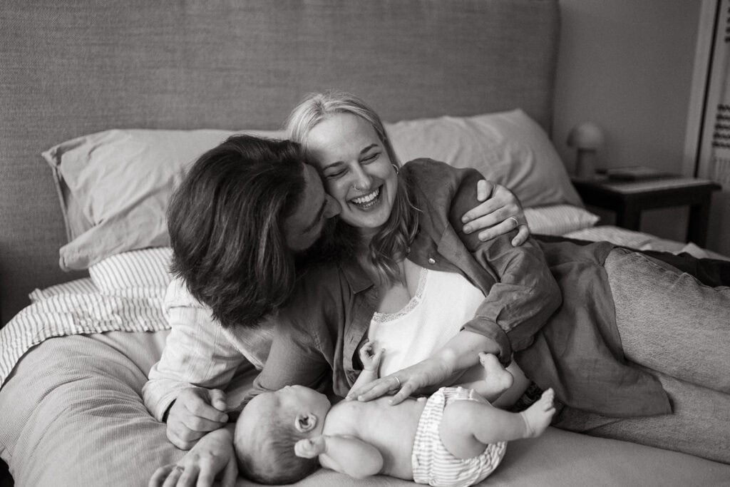 a man hugs his laughing partner they are on a bed near their new baby