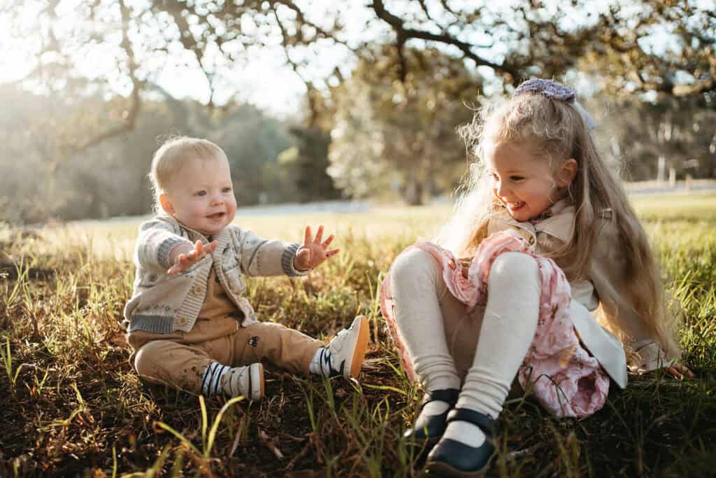 two little kids play on the grass they are sitting with knees up and hands out