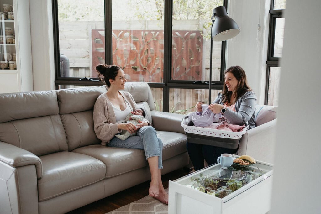 a melbourne postpartum doula talks with a new mother while folding laundry, the mother is holding her newborn baby