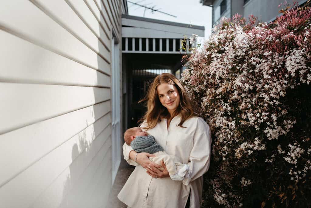 mother holding her newborn baby at a melbourne home in the sunshine near a flowering plant